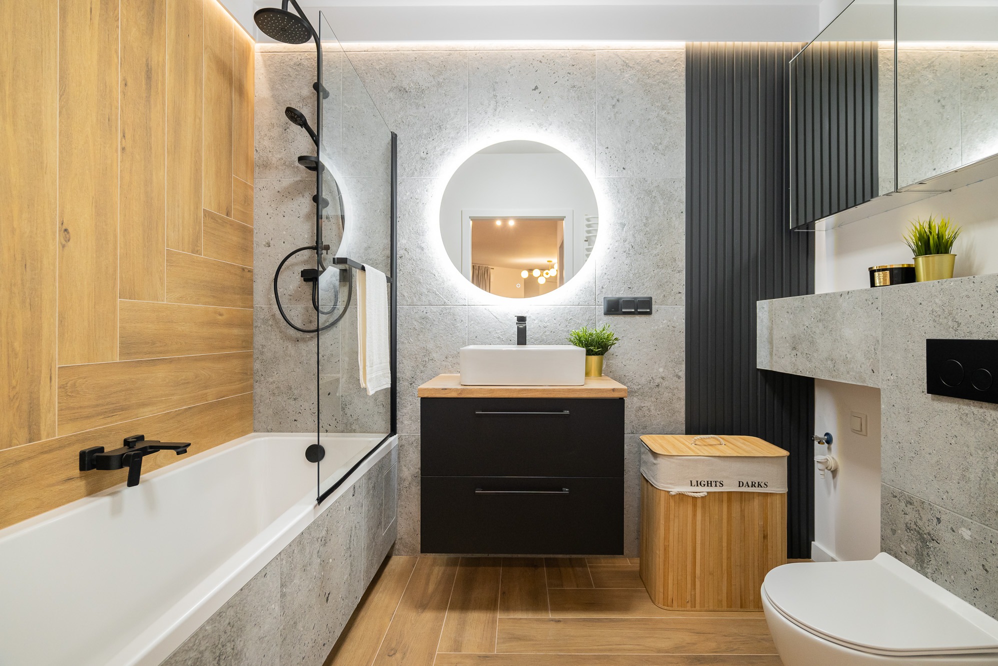Nowoczesna łazienka z drewnopodobnymi i szarymi płytkami, okrągłym lustrem z podświetleniem LED, czarną armaturą i drewnianym koszem na pranie z napisem 'LIGHTS DARKS'.