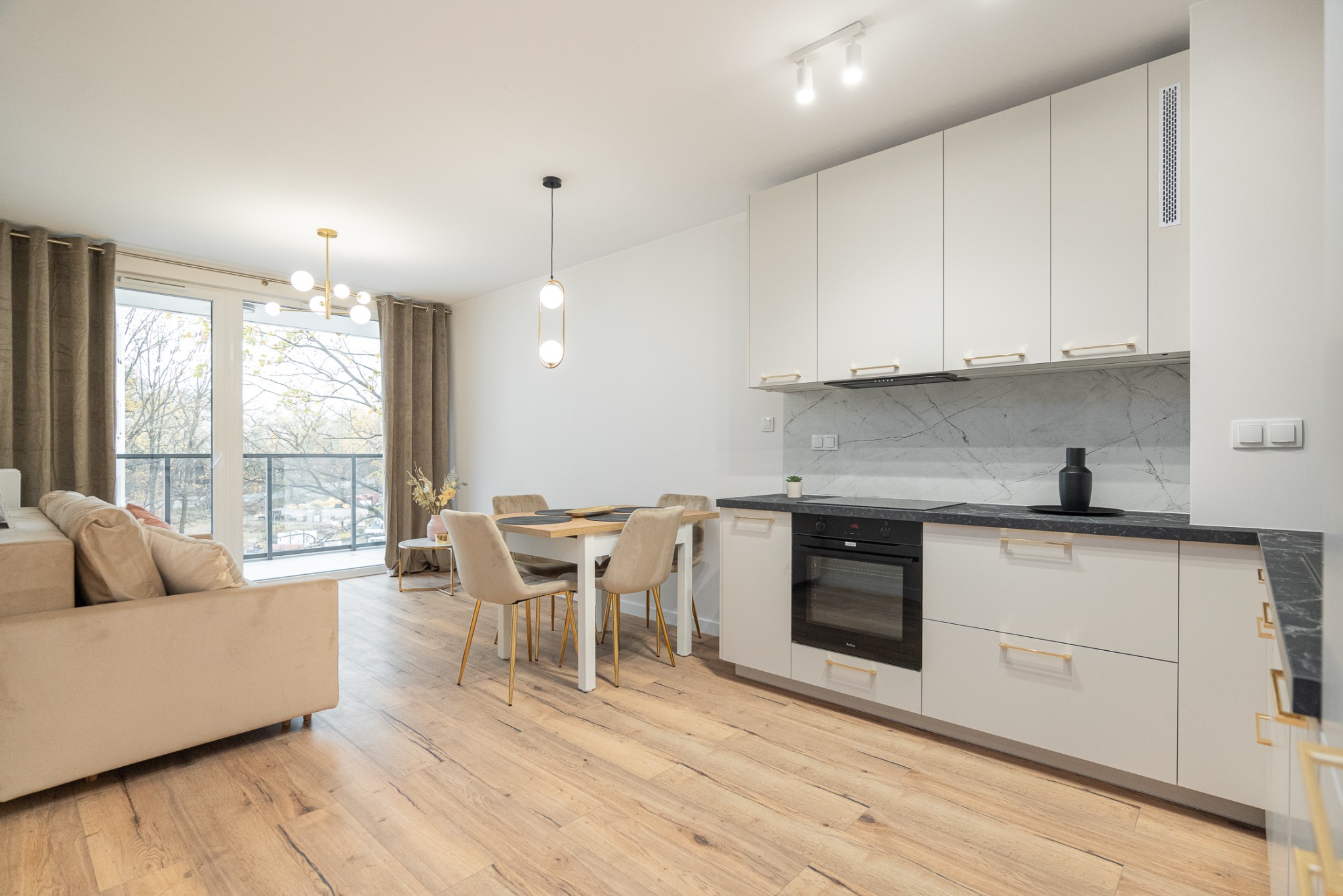 Jasne wnętrze mieszkania z drewnianą podłogą, białymi meblami kuchennymi z marmurowym blatem i złotymi uchwytami, oraz częścią wypoczynkową z beżową sofą i balkonem z widokiem na drzewa.