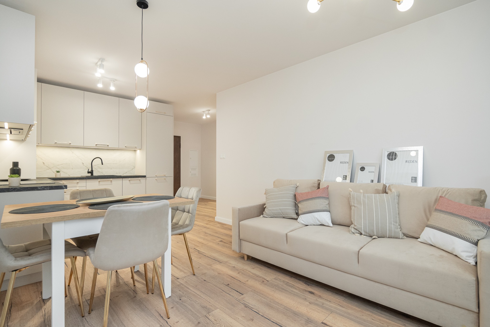 Jasne wnętrze mieszkania z drewnianą podłogą, beżową sofą z dekoracyjnymi poduszkami, stołem z krzesłami na złotych nogach oraz kuchnią w tle z czarnymi akcentami.