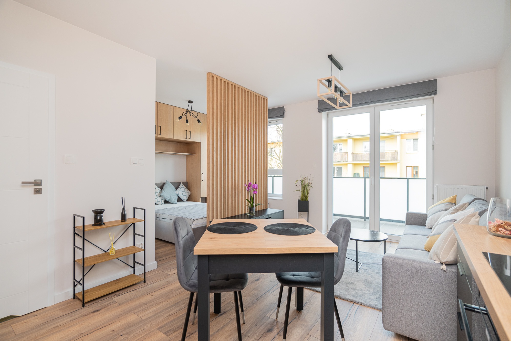 Jasne wnętrze mieszkania z drewnianą podłogą, stołem jadalnym z szarymi krzesłami, szarą sofą przy balkonie i drewnianym regałem na dekoracje, oddzielone ażurową ścianką od sypialni z łóżkiem.