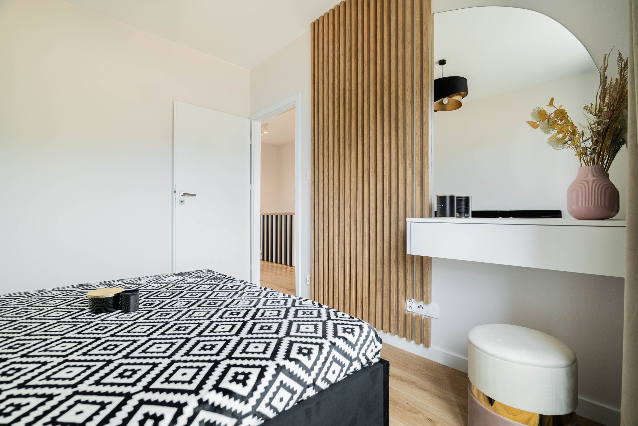 Nowoczesna sypialnia z drewnianą ścianką działową, toaletką z owalnym lustrem i geometryczną pościelą w czarno-białe wzory.
