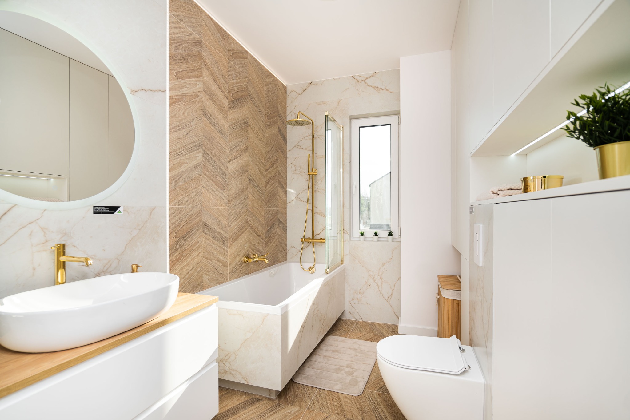 Nowoczesna łazienka z wanną obudowaną marmurem, złotą armaturą, umywalką na drewnianym blacie i ścianą w jodełkę z płytek imitujących drewno.
