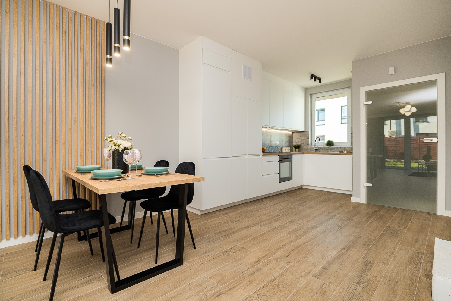 Jasna kuchnia z drewnopodobną podłogą, białymi szafkami, drewnianym stołem z czarnymi krzesłami i dekoracyjną ścianą z drewnianych listew.