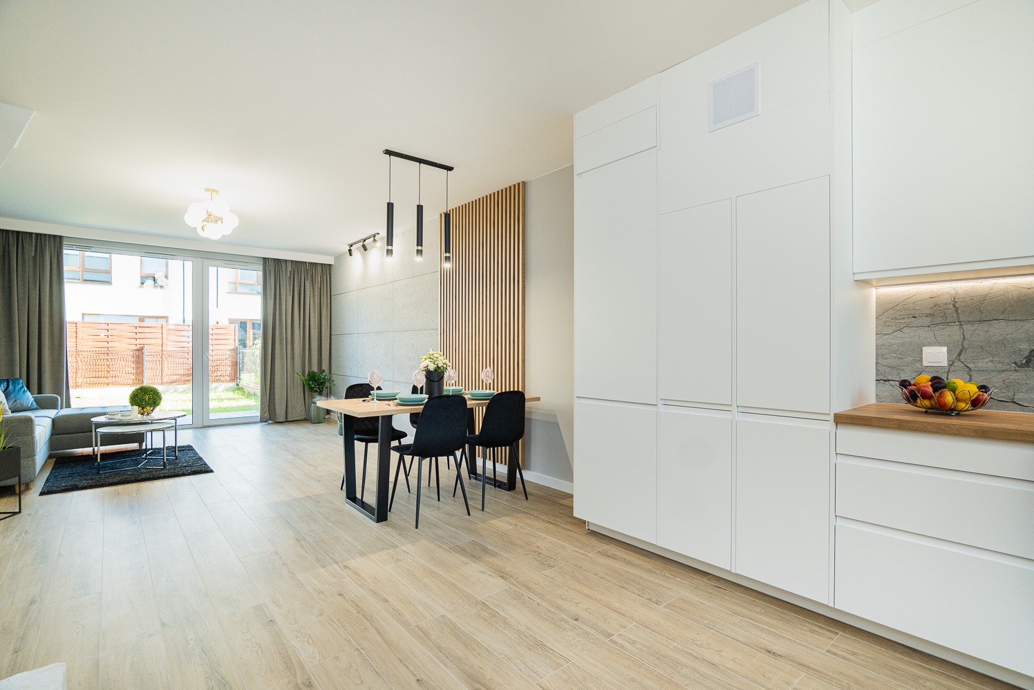 Jasne, przestronne wnętrze z drewnianą podłogą, stołem z czarnymi krzesłami i białymi meblami kuchennymi, oświetlone wiszącymi lampami i światłem dziennym wpadającym przez duże okno tarasowe.