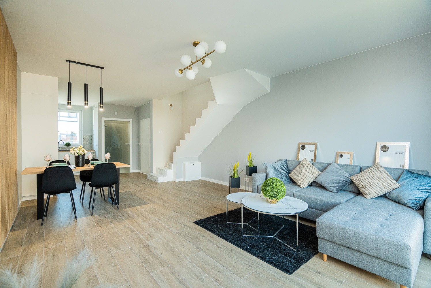 Jasne wnętrze z drewnopodobną podłogą, szarą sofą narożną, białymi schodami i stołem jadalnym z czarnymi krzesłami, oświetlone nowoczesnymi lampami.