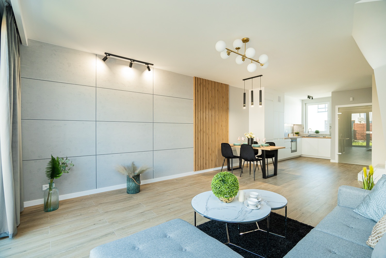 Jasne, przestronne wnętrze salonu z aneksem kuchennym, ściana z szarych paneli, drewniane lamele, parkiet ułożony w jodełkę, nowoczesne oświetlenie punktowe i wiszące, szara sofa i marmurowe...