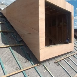 Wladyslaw Byczek - Surowa konstrukcja okna dachowego z drewna na pokrytym papą dachu, widoczne krokwie i narzędzia wewnątrz otworu okiennego, błękitne niebo w tle.