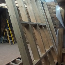 Wladyslaw Byczek - Konstrukcja szkieletowa z drewna, widoczne elementy pionowe i poziome, w tle drabina i ceglana ściana, surowe wnętrze w trakcie budowy.