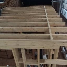 Wladyslaw Byczek - Drewniany szkielet podłogi z belek, widoczne fragmenty ceglanej ściany i rusztowania, w tle ciemna przestrzeń strychu.