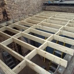 Wladyslaw Byczek - Drewniana konstrukcja szkieletowa pod podłogę z widoczną ceglaną ścianą w tle i żółtą drabinką, etap przygotowawczy przed ułożeniem warstwy wierzchniej.