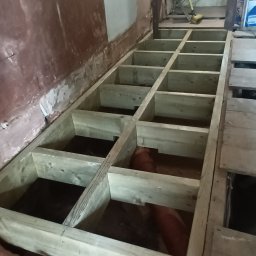 Wladyslaw Byczek - Konstrukcja podłogi z drewnianych belek, widoczne rury instalacji w przestrzeni między belkami, surowa ściana w tle.