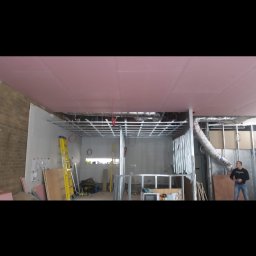 Wladyslaw Byczek - Wnętrze w trakcie prac wykończeniowych, widoczny metalowy stelaż sufitu podwieszanego z zamontowanymi płytami gipsowo-kartonowymi w kolorze różowym, drabina i materiały budowlane w tle,...