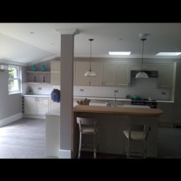 Wladyslaw Byczek - Jasna kuchnia z wyspą i dwoma chromowanymi krzesłami, widoczne płytki na ścianie, okno i dekoracje na półkach.