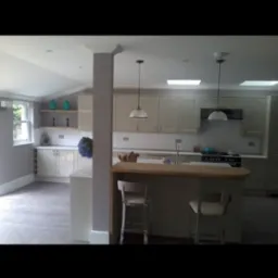 Jasna kuchnia z wyspą i dwoma chromowanymi krzesłami, widoczne płytki na ścianie, okno i dekoracje na półkach.