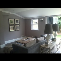 Wladyslaw Byczek - Jasne, nowocześnie urządzone wnętrze salonu z szarą sofą, drewnianym stolikiem kawowym i dekoracyjnymi lampami, widok na ogród przez przeszklone drzwi balkonowe.