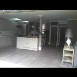 Przestronne wnętrze domu z otwartą kuchnią i salonem, wykończone w jasnych barwach, z podłogą imitującą drewno i wyspą kuchenną.