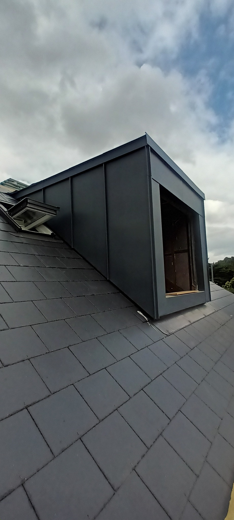 Nowa, grafitowa lukarna z metalowym obiciem na dachu pokrytym ciemną dachówką, widok z dołu na pochmurne niebo.