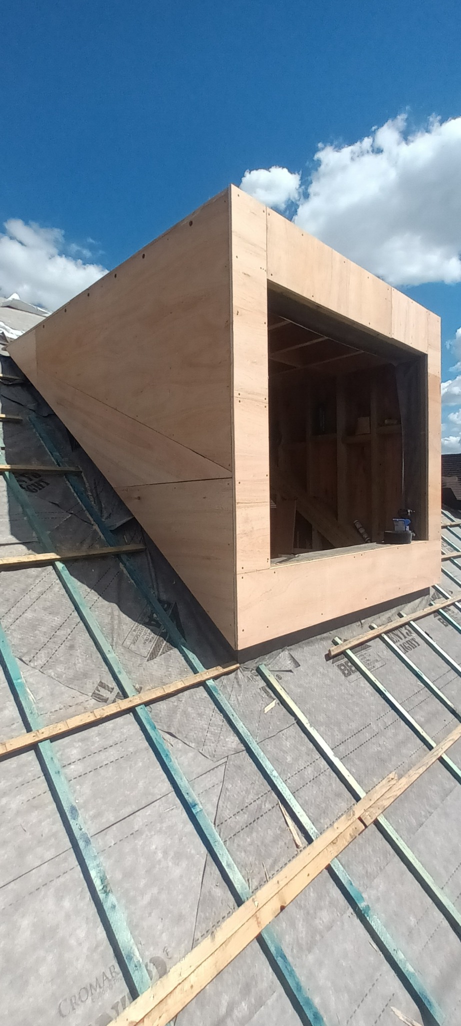 Surowa konstrukcja okna dachowego z drewna na pokrytym papą dachu, widoczne krokwie i narzędzia wewnątrz otworu okiennego, błękitne niebo w tle.