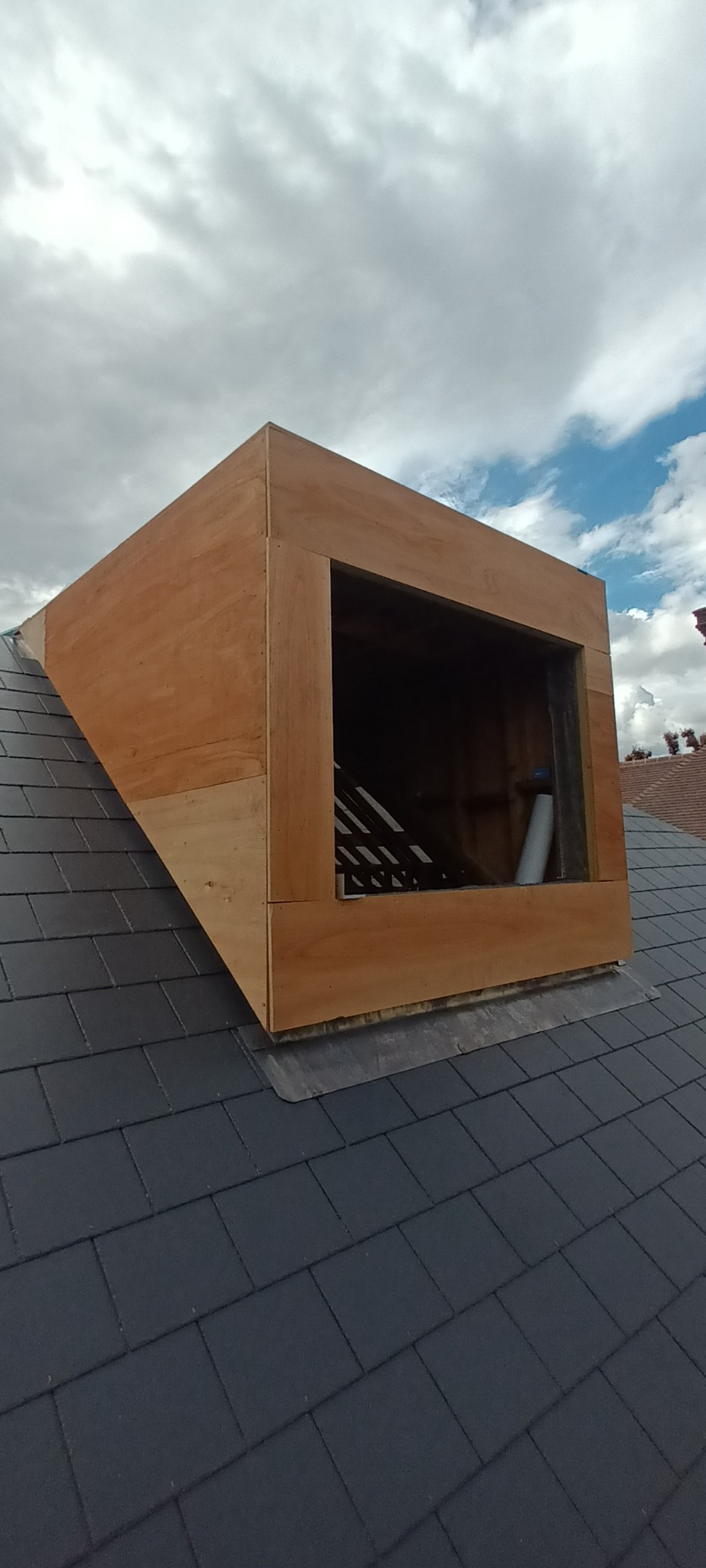 Niedokończona drewniana konstrukcja okna dachowego osadzona na pokrytym dachówką dachu, widoczne elementy więźby dachowej wewnątrz otworu okiennego, pochmurne niebo w tle.