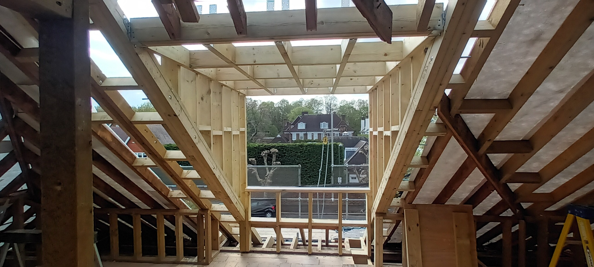 Konstrukcja drewniana poddasza z widokiem na dom i zieleń za oknem, etap budowy widoczny od wewnątrz.