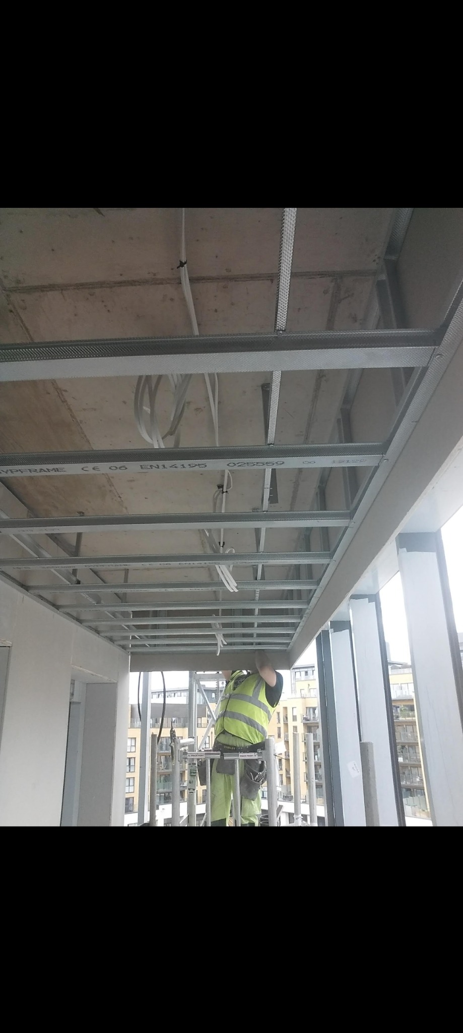 Pracownik w kamizelce odblaskowej montuje metalowy ruszt sufitowy w nowym budynku, widoczne przewody i elementy konstrukcyjne, w tle okna z widokiem na zabudowę miejską.