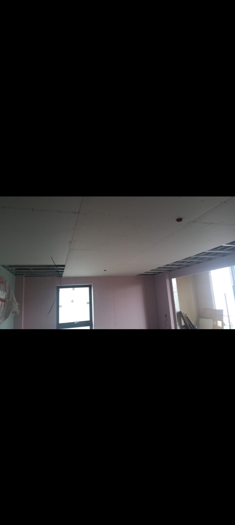 Montaż sufitu podwieszanego z płyt kartonowo-gipsowych, widoczna metalowa konstrukcja nośna, częściowo zamontowane płyty, ściany przygotowane pod malowanie na różowo, okno w tle.