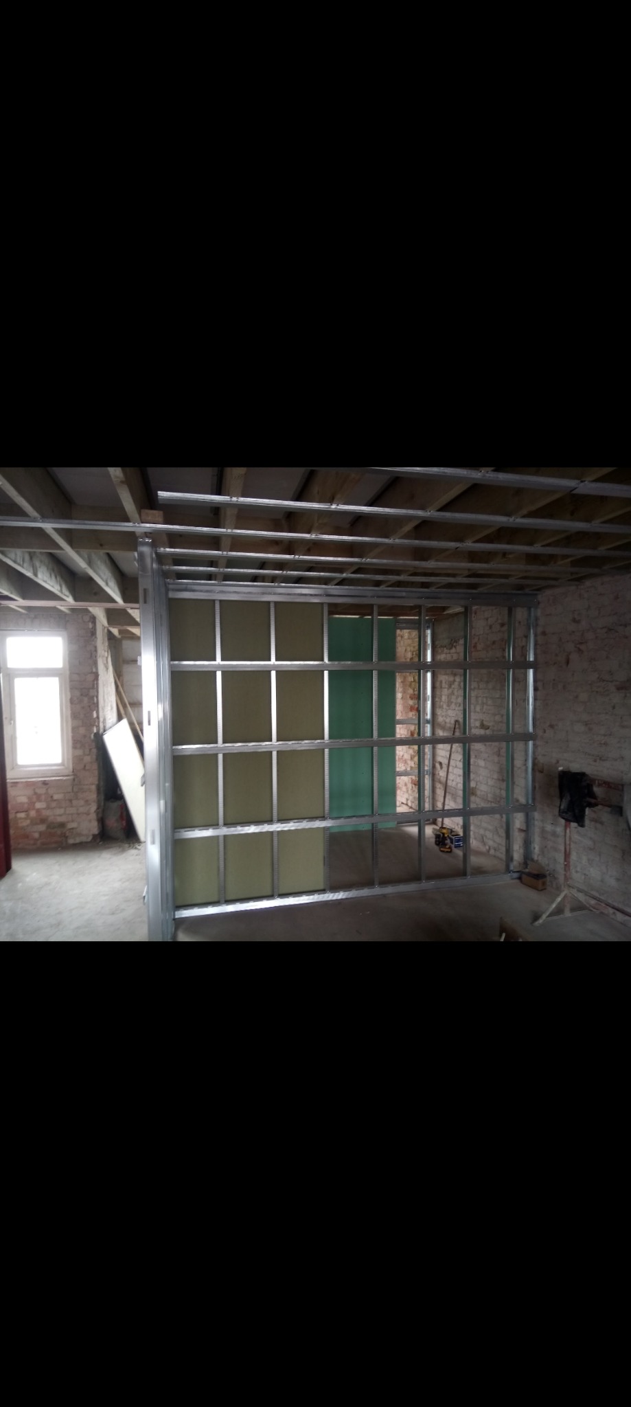 Montaż metalowej konstrukcji szkieletowej z częściowo zamontowaną płytą gipsowo-kartonową w surowym wnętrzu z ceglanymi ścianami i drewnianym stropem.