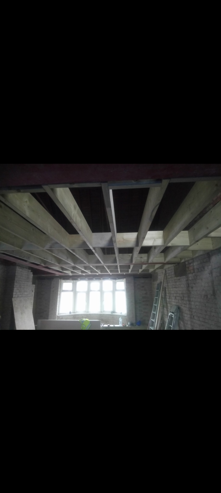 Drewniana konstrukcja sufitu podwieszanego w trakcie montażu, widoczne belki i stelaż na tle okna i surowych ścian z cegły, drabina oparta o ścianę.