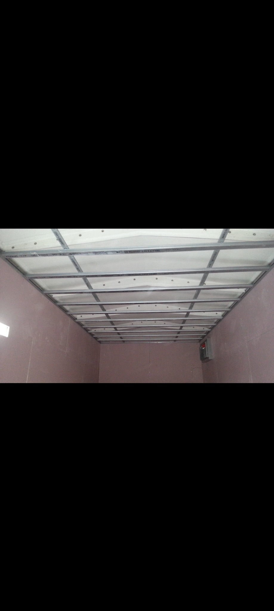 Sufit z płyt karton gips, widoczne metalowe profile konstrukcyjne i białe płyty, ściany w kolorze pudrowego różu, etap montażu.