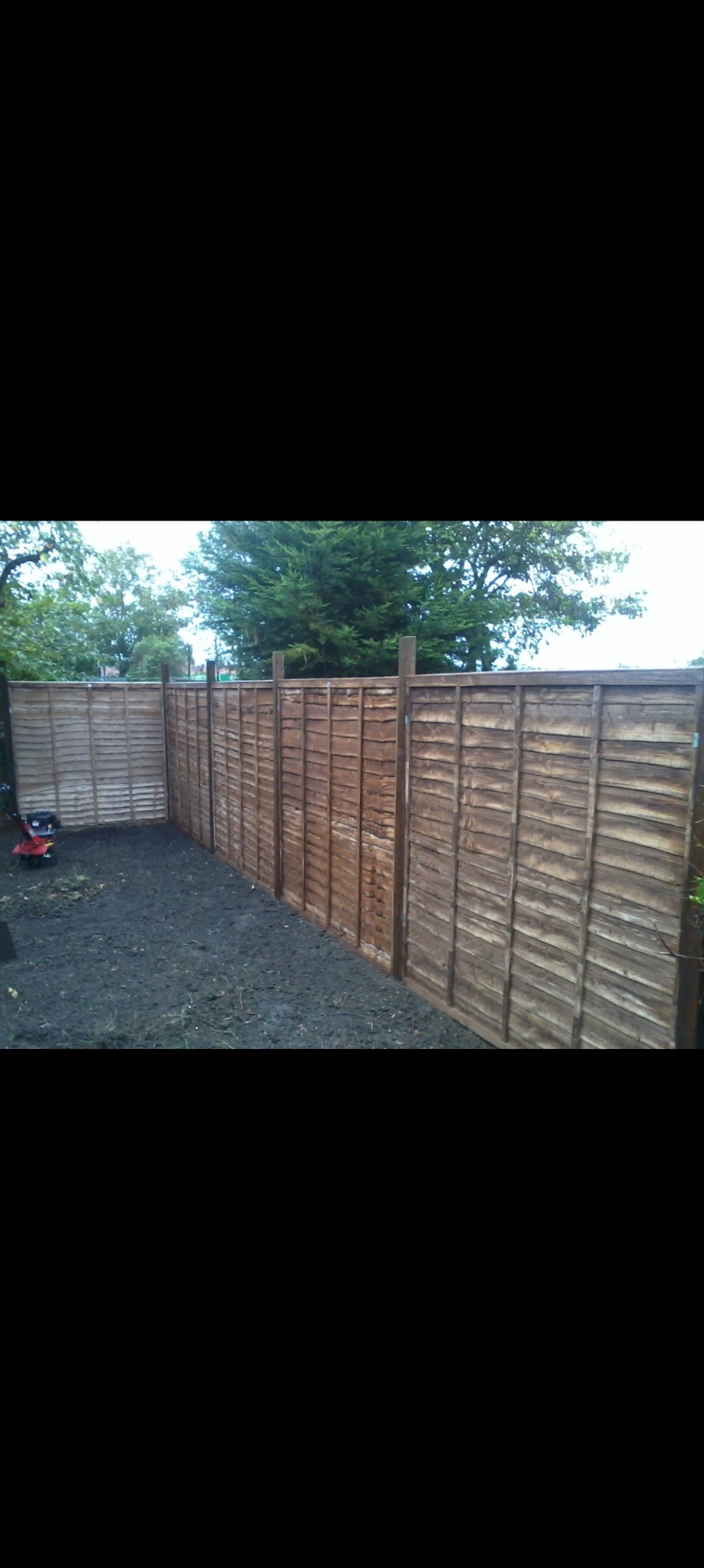 Nowo postawione drewniane ogrodzenie panelowe z widoczną glebą i fragmentem glebogryzarki w tle.