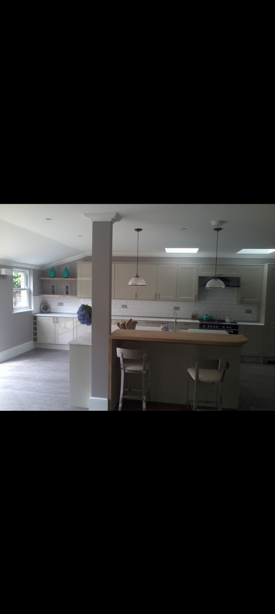 Jasna kuchnia z wyspą i dwoma chromowanymi krzesłami, widoczne płytki na ścianie, okno i dekoracje na półkach.