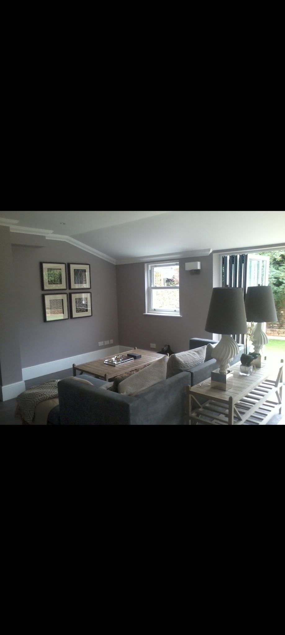 Jasne, nowocześnie urządzone wnętrze salonu z szarą sofą, drewnianym stolikiem kawowym i dekoracyjnymi lampami, widok na ogród przez przeszklone drzwi balkonowe.