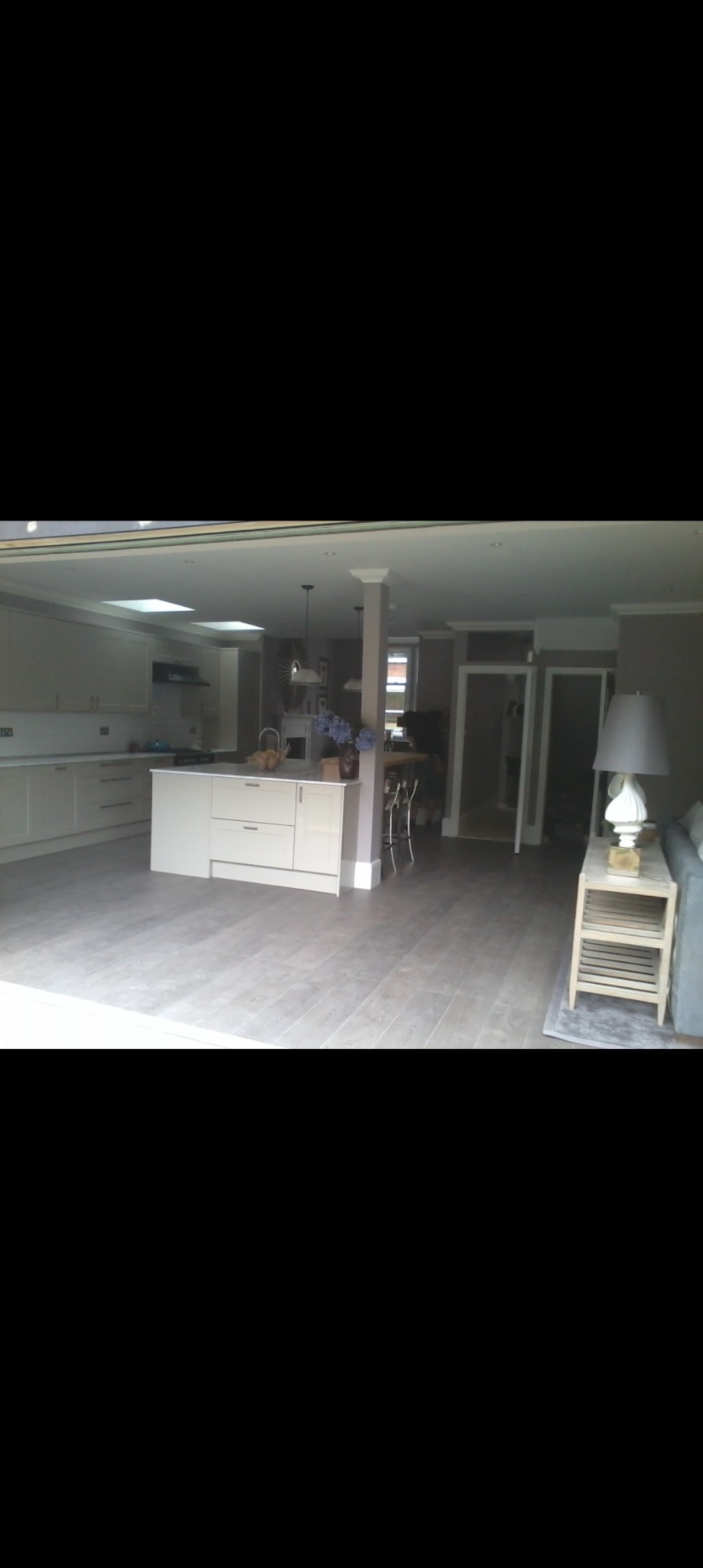 Przestronne wnętrze domu z otwartą kuchnią i salonem, wykończone w jasnych barwach, z podłogą imitującą drewno i wyspą kuchenną.