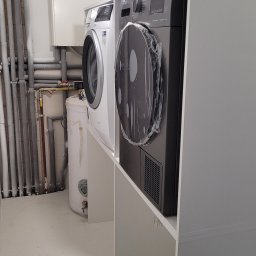 Efneo Fryderyk Migaszewski - Pralka i suszarka ustawione na białym stelażu w pomieszczeniu gospodarczym z instalacją wodną i grzewczą na ścianie, suszarka jeszcze w folii ochronnej.