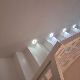 Efneo Fryderyk Migaszewski - Nowoczesne, białe schody z mikrobetonu oświetlone punktowymi lampami w suficie, widok od dołu, z ozdobną, ażurową balustradą.