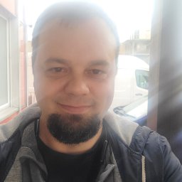 Efneo Fryderyk Migaszewski - Portret mężczyzny z brodą, ubranego w bluzę z kapturem i kurtkę, na tle częściowo widocznego białego samochodu dostawczego i budynku.