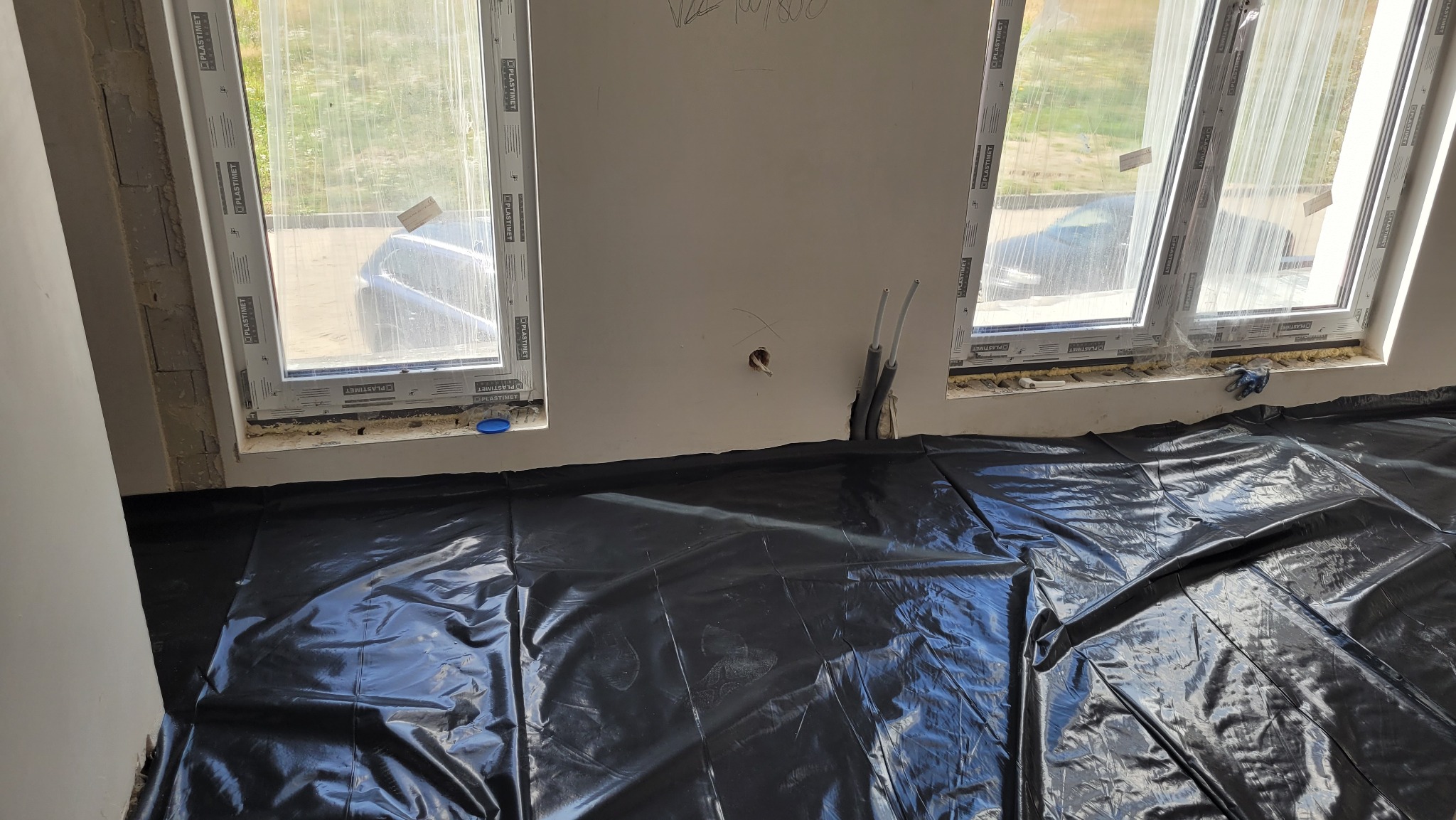Przygotowanie podłoża pod wylewkę: czarna folia zabezpieczająca rozłożona na podłodze w pomieszczeniu z oknami, widoczne rury instalacji wystające ze ściany, okna uszczelnione pianką montażową.