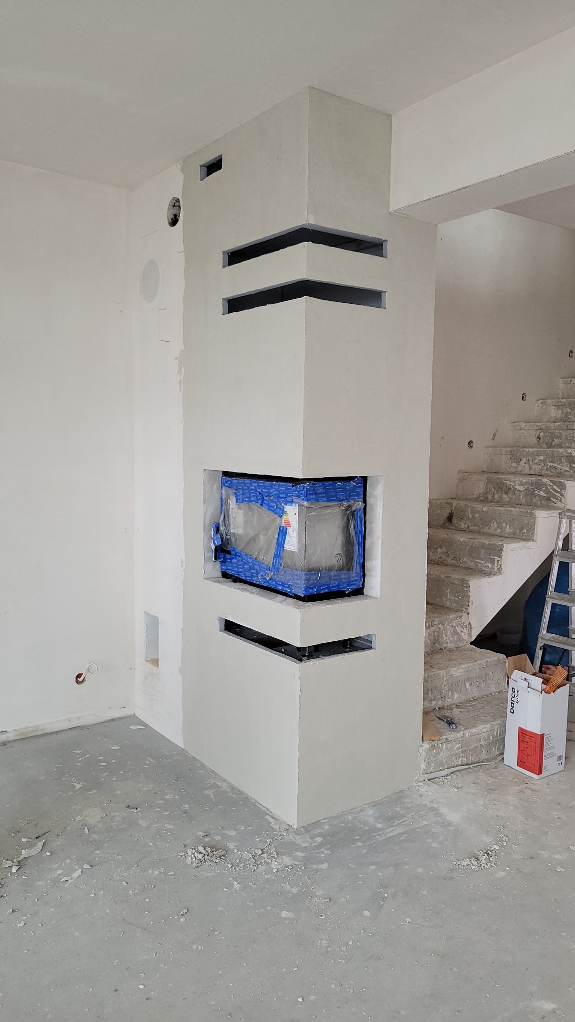 Wnętrze w trakcie prac wykończeniowych: obudowa kominka z otworami wentylacyjnymi, zabezpieczony kominek w folii, surowe schody betonowe w tle.