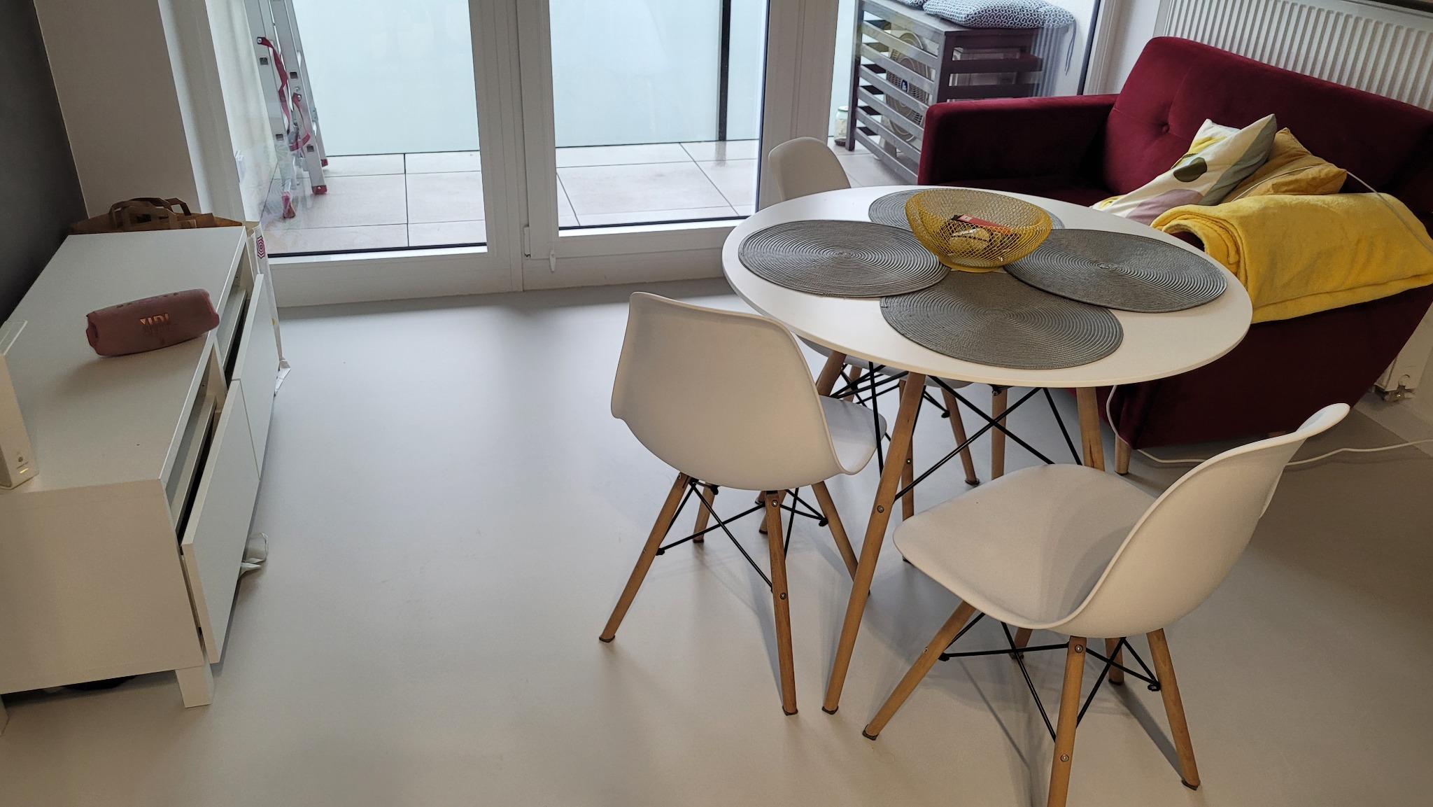 Jasna podłoga z żywicy w nowoczesnym mieszkaniu, widoczny okrągły stół z szarymi podkładkami, białe krzesła, czerwona sofa z żółtym kocem oraz biała szafka RTV z głośnikiem JBL.