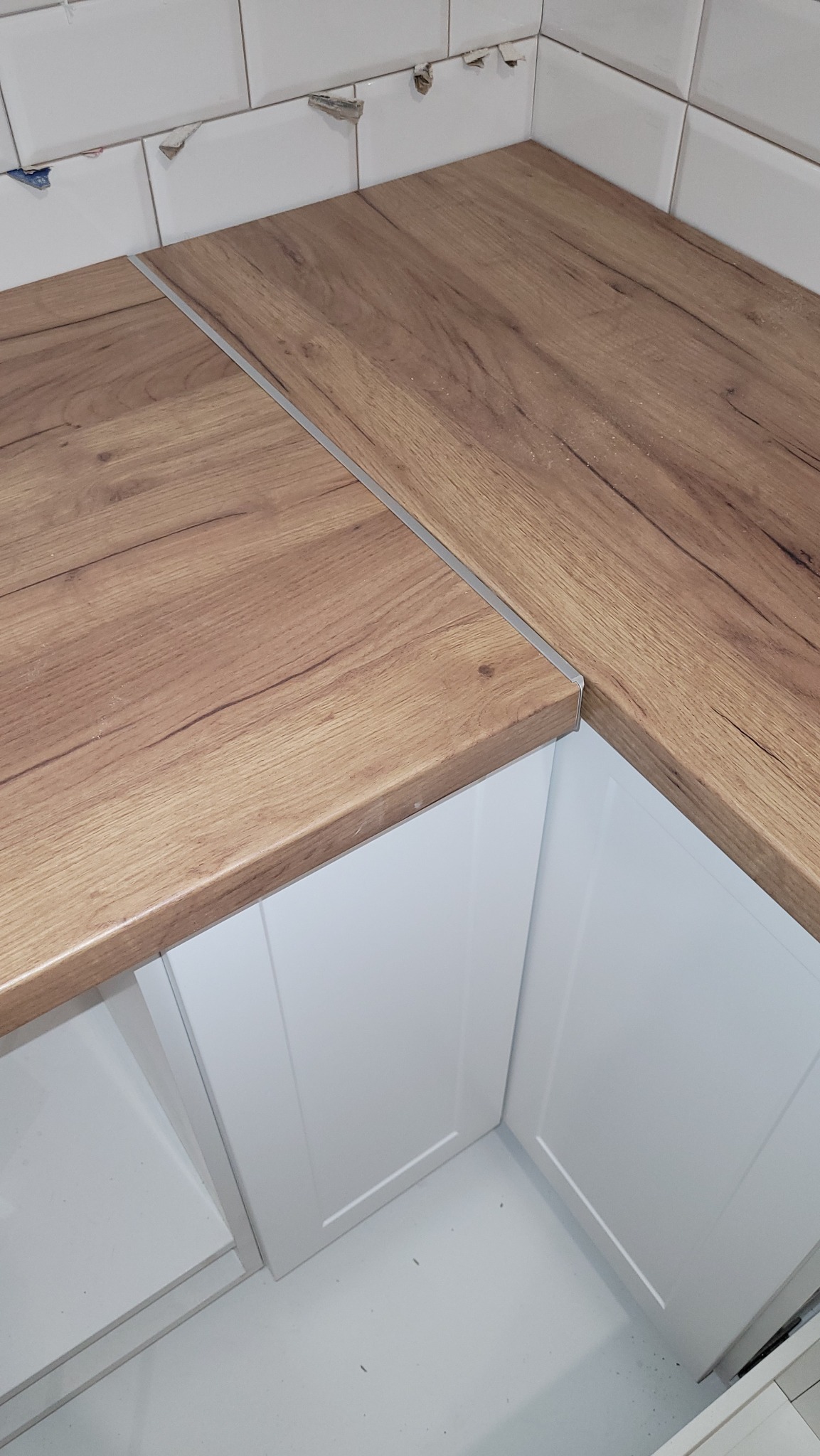 Fragment narożnej zabudowy kuchennej z drewnianym blatem i białymi szafkami, widoczne niedokończone białe płytki na ścianie, brak fugi.