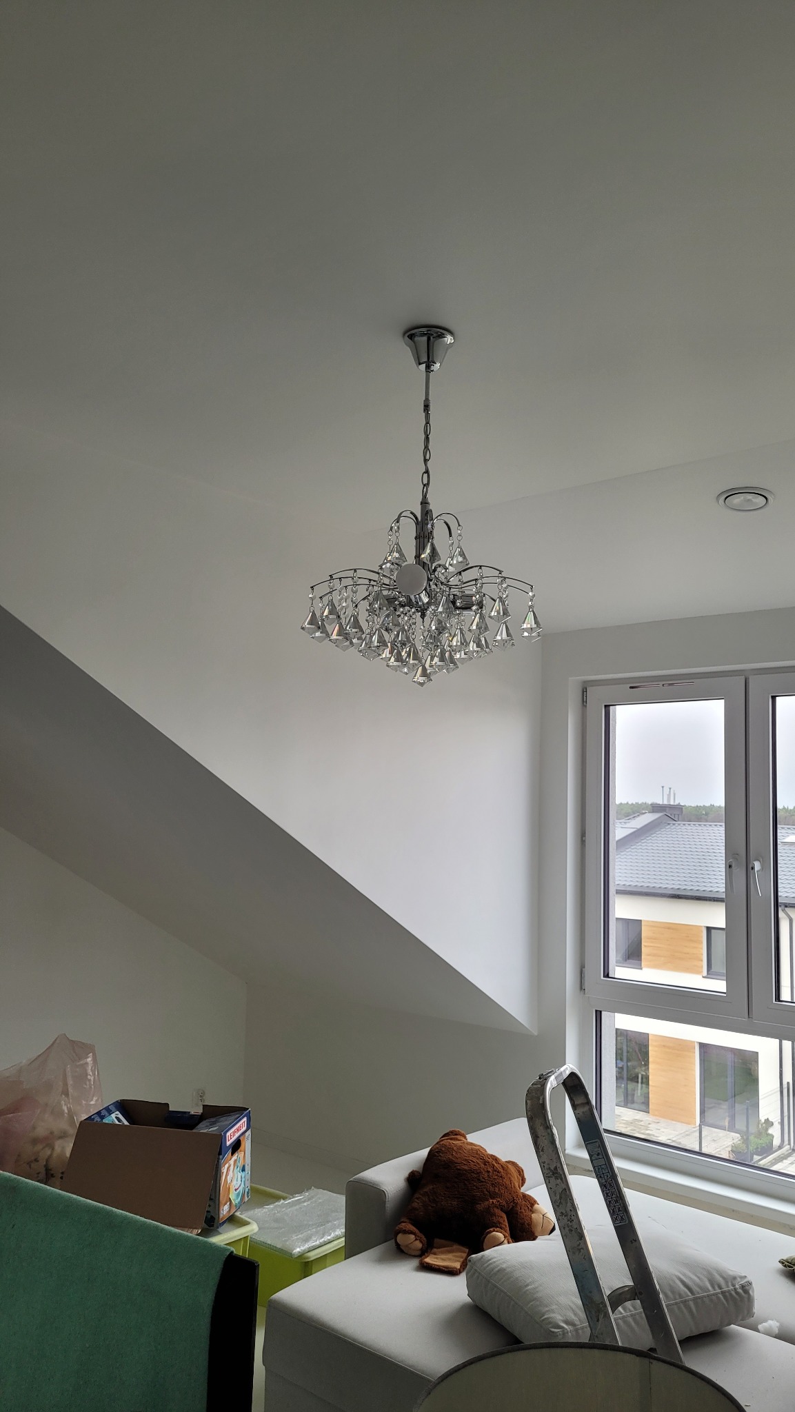 Kryształowy żyrandol zawieszony na suficie w pokoju na poddaszu, widoczne skosy, okno z widokiem na domy, na kanapie leży pluszowy miś i oparta jest drabina
