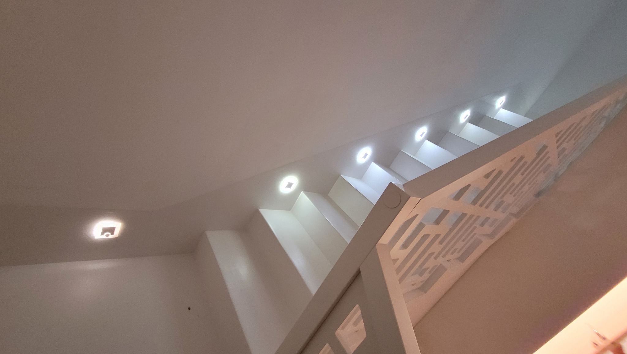 Nowoczesne, białe schody z mikrobetonu oświetlone punktowymi lampami w suficie, widok od dołu, z ozdobną, ażurową balustradą.