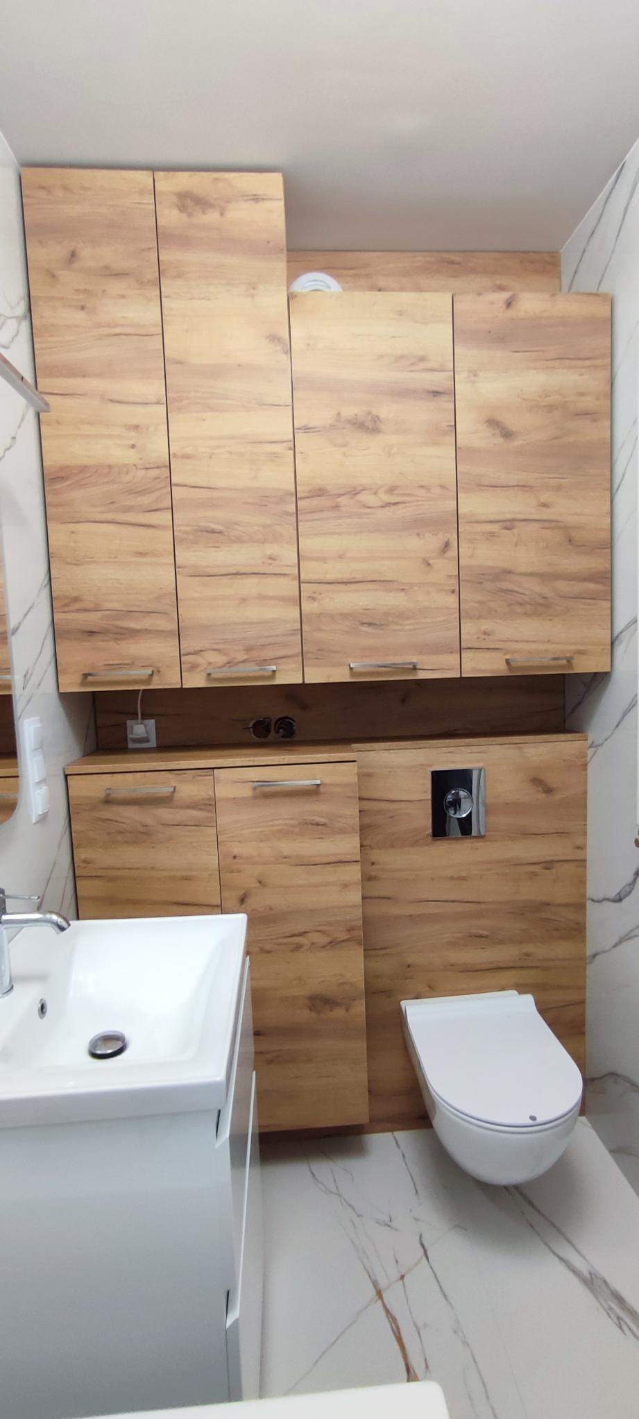 Nowoczesna łazienka z zabudową z drewnopodobnej płyty, wisząca toaleta, umywalka nablatowa i marmurowe płytki podłogowe.
