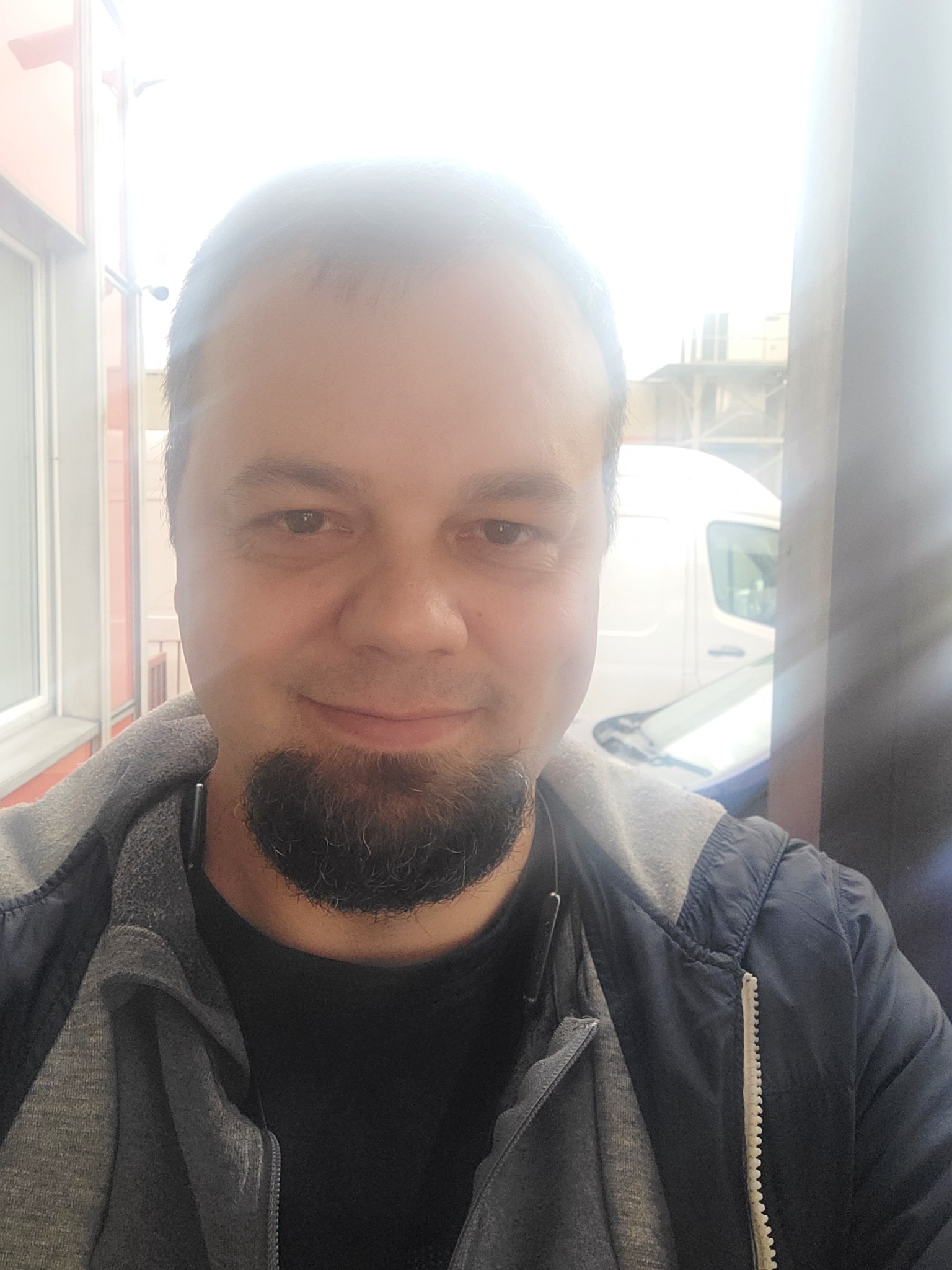 Portret mężczyzny z brodą, ubranego w bluzę z kapturem i kurtkę, na tle częściowo widocznego białego samochodu dostawczego i budynku.