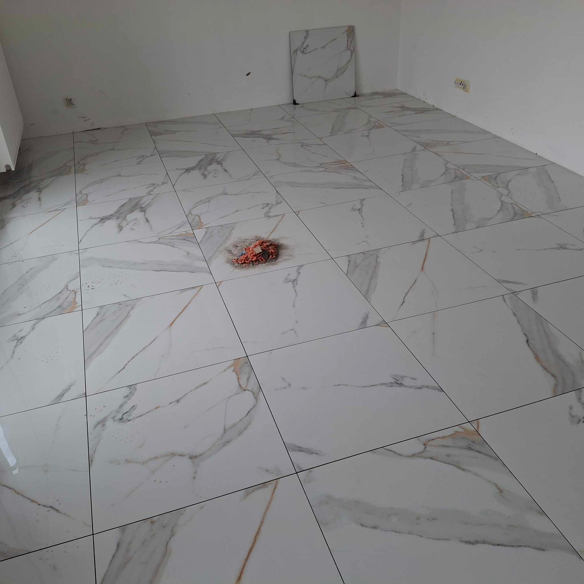 Ułożone, białe płytki podłogowe z szarym i brązowym wzorem żyłkowania, niedbale pozostawiona szmatka na środku, ślady po fugowaniu na powierzchni płytek, gniazdko elektryczne na ścianie i dodatkowa...