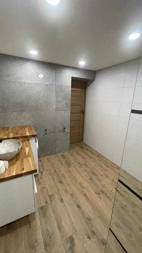 Nowoczesna łazienka z drewnopodobną podłogą, szarymi płytkami i drewnianym blatem pod umywalkę, drzwi w tle.