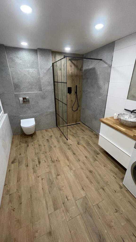 Nowoczesna łazienka z drewnopodobną podłogą, szarą płytką imitującą beton na ścianach, kabiną prysznicową z czarnymi szprosami i umywalką nablatową w stylu rustykalnym.