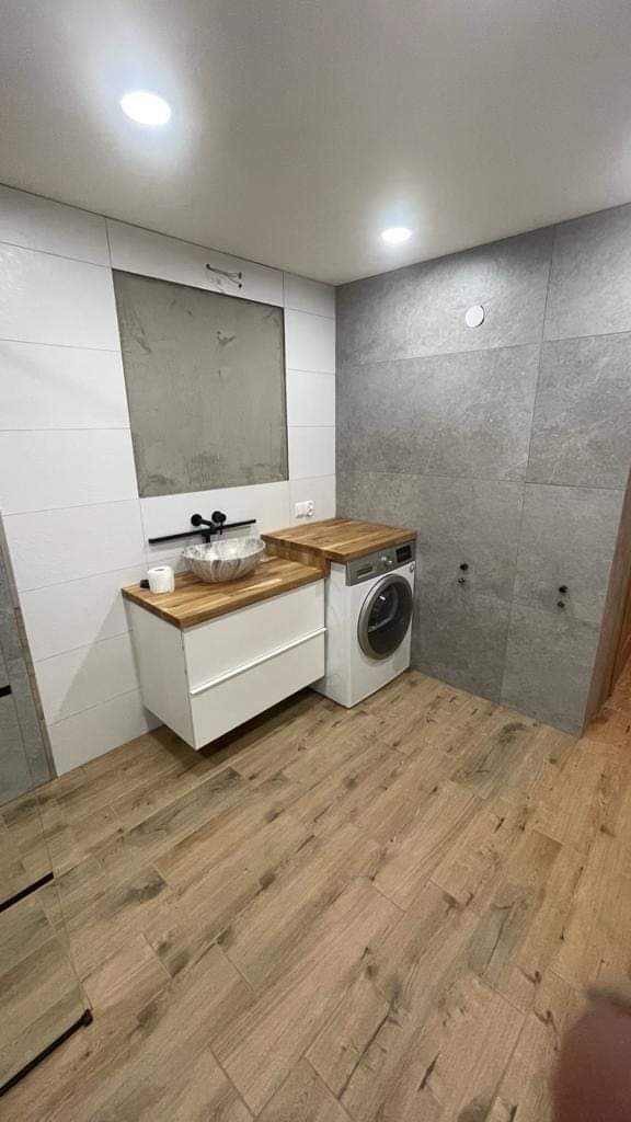 Nowoczesna łazienka w trakcie wykończenia, z drewnianym blatem pod umywalką i pralką, szare płytki na ścianach, jasne płytki podłogowe imitujące drewno, widoczne niedokończone miejsce na lustro.