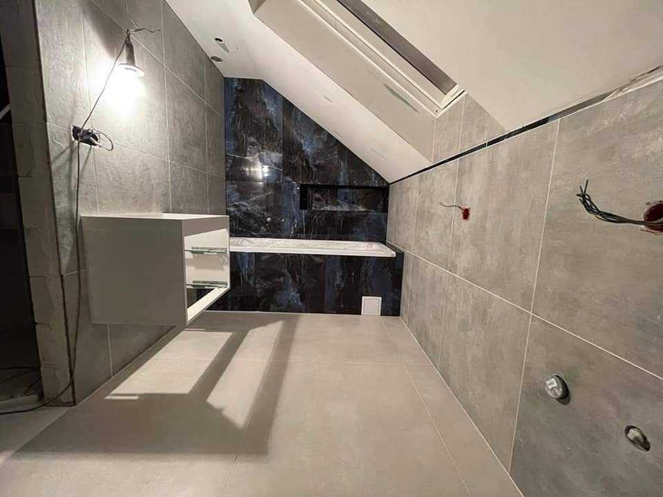 Wnętrze łazienki w trakcie wykańczania, widoczne szare płytki na ścianach i podłodze, czarne płytki imitujące marmur przy wannie, biała szafka pod umywalkę, wystające kable i otwory na oświetlenie.