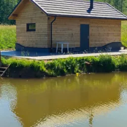 Drewniany domek letniskowy z ciemną stolarką okienną i kominem, posadowiony nad brzegiem stawu, otoczony zielenią.